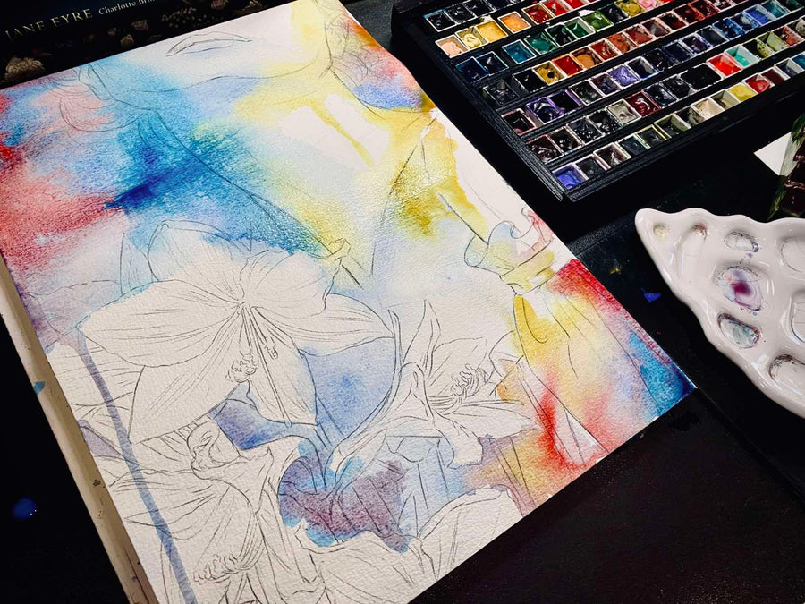 Artistic watercolor painting of flowers on a canvas with watercolor pans and a palette in the background.